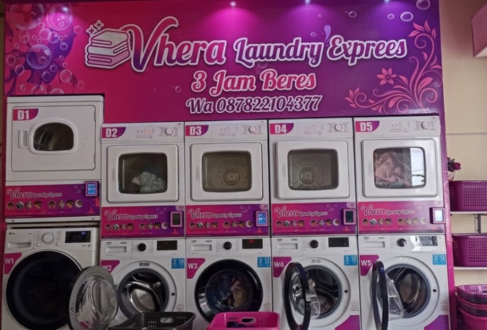Deretan mesin cuci dan pengering di Vhera Laundry Express dengan latar belakang berwarna merah muda.